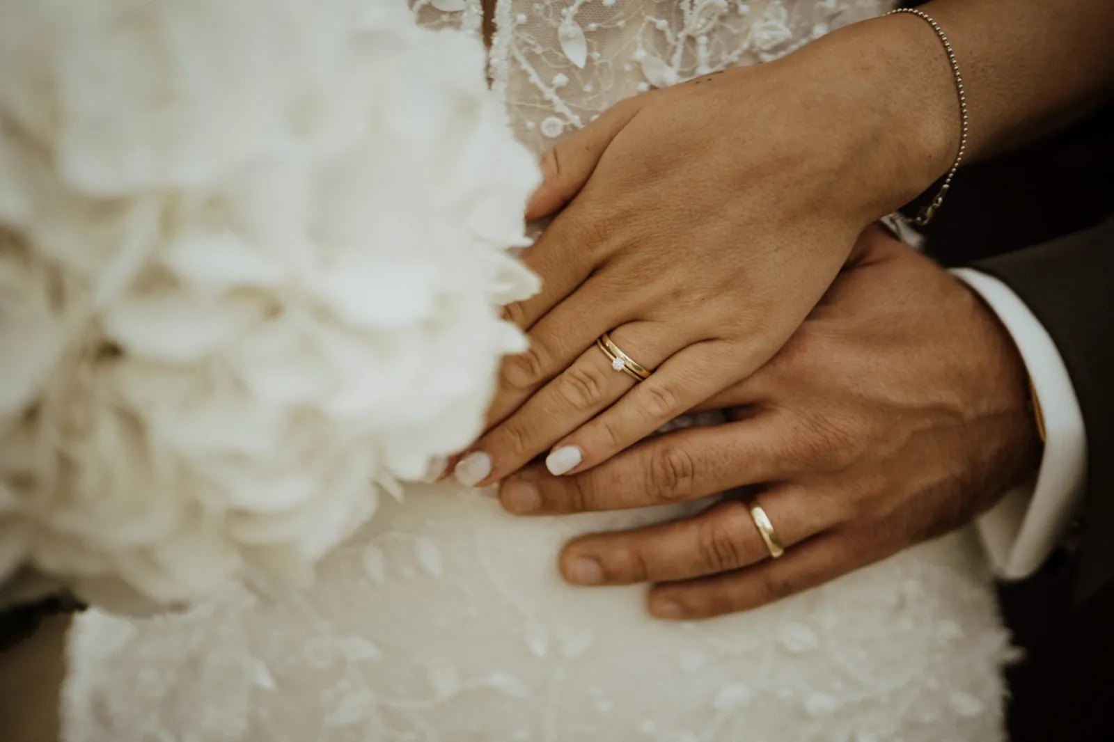 Luisa & Norman – Die Hände von Braut und Bräutigam mit Eheringen liegen auf dem weißen Hochzeitskleid