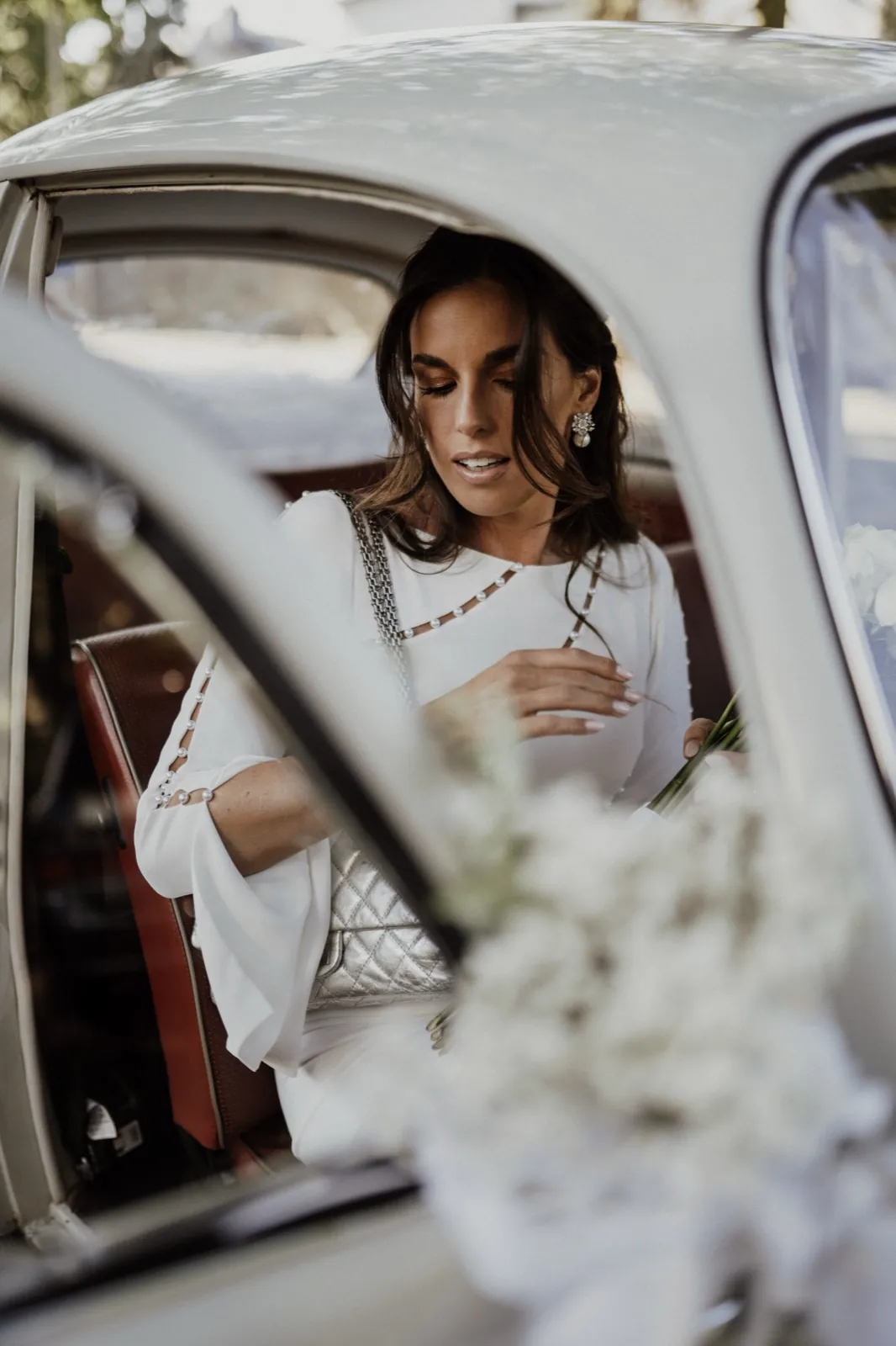 Clarissa & Kai – Eine Braut im weißen Kleid sitzt in einem weißen Oldtimer und hält einen Blumenstrauß