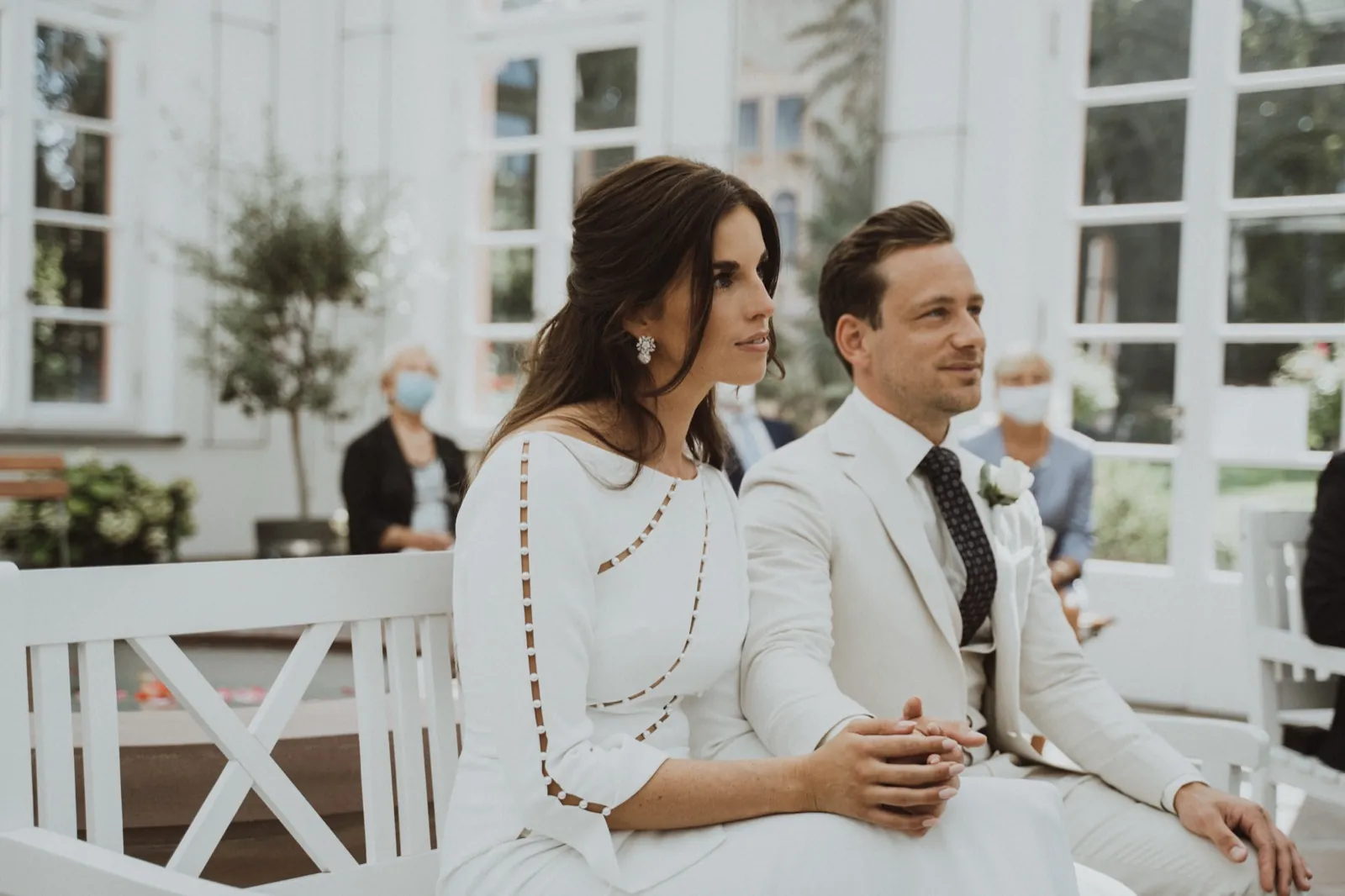 Clarissa & Kai – Braut und Bräutigam sitzen während der Trauung Hand in Hand auf einer weißen Bank