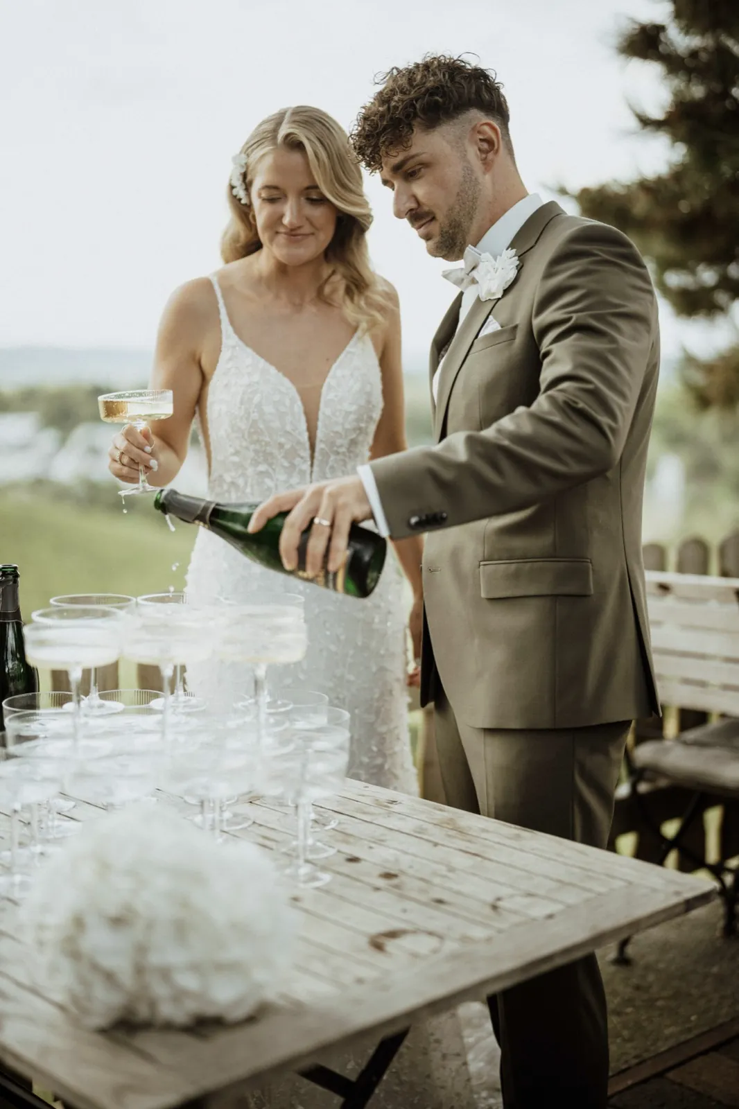Luisa & Norman – Ein Brautpaar gießt Champagner in eine Pyramide aus Gläsern auf einem Holztisch