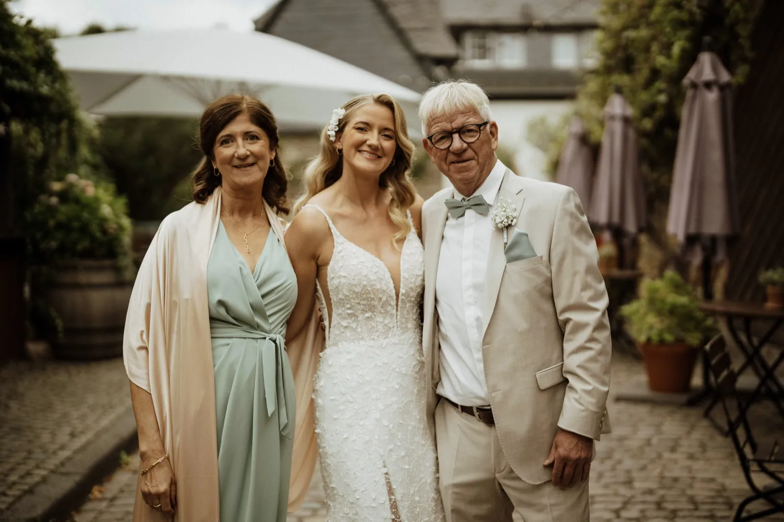 Luisa & Norman – Eine Braut steht lächelnd zwischen ihren Eltern im Außenbereich einer Hochzeitslocation