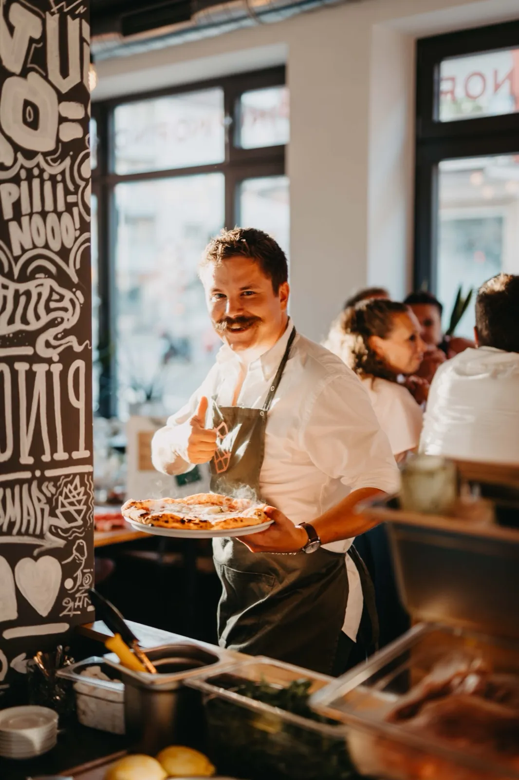 Amelie & Andi – Ein lächelnder Koch mit Schnurrbart präsentiert eine frische Pizza in einem Restaurant