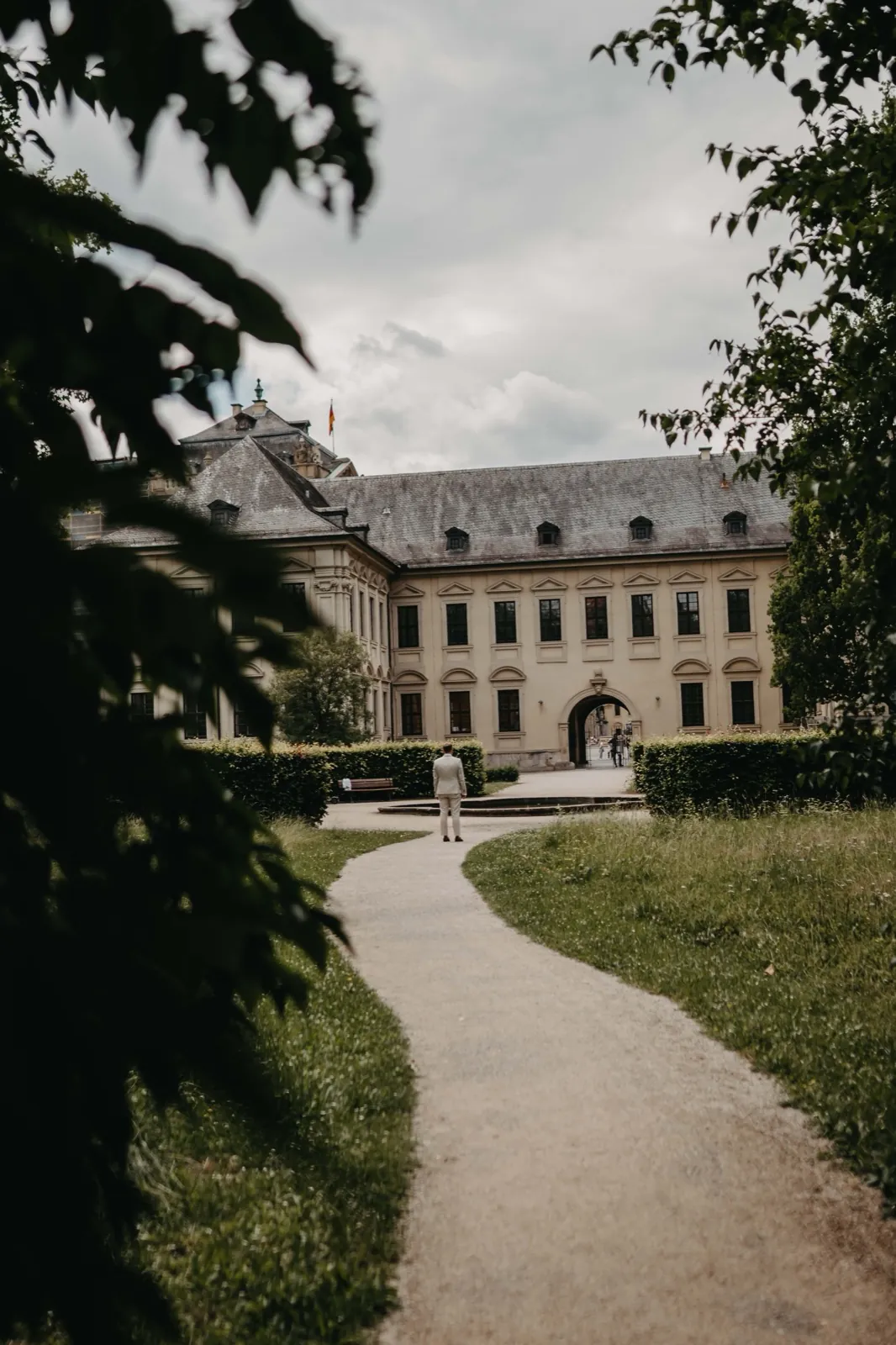 Amelie & Andi – Ein Mann im hellen Anzug steht auf einem Weg vor einem großen Schloss