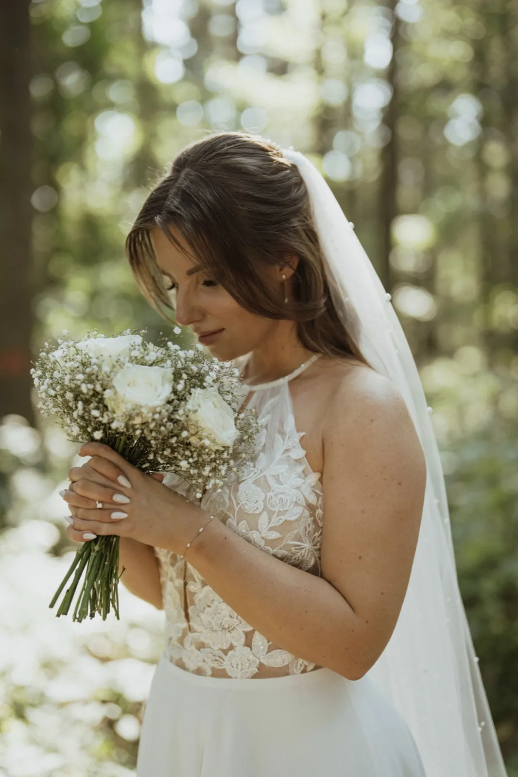 Anna & Moritz – Eine Braut im weißen Kleid blickt im Wald lächelnd auf ihren Brautstrauß