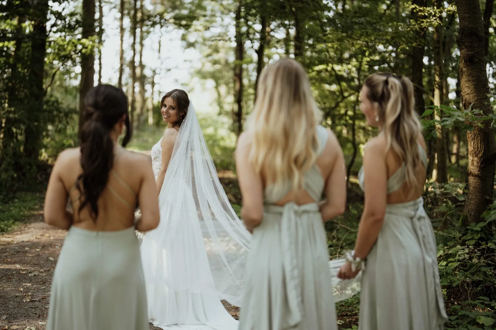 Anna & Moritz – Eine Braut blickt im Wald lächelnd zurück zu ihren drei Brautjungfern