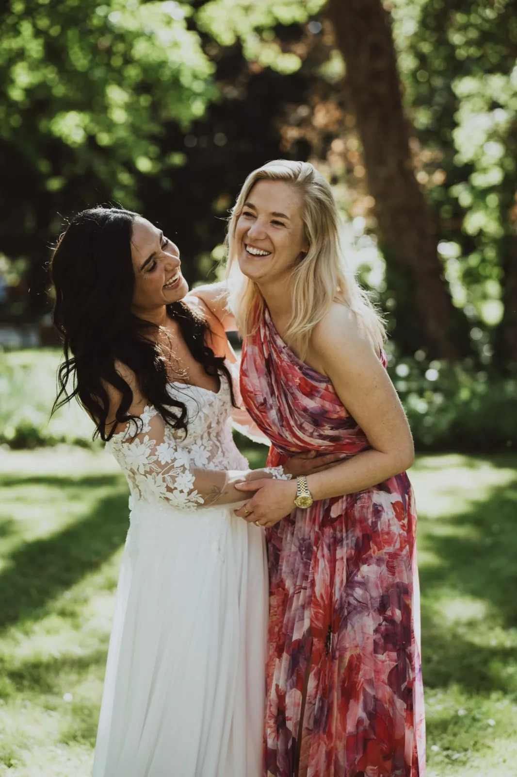 Caro & Dominik – Eine Braut im weißen Kleid lacht herzlich mit einer Freundin im bunten Kleid