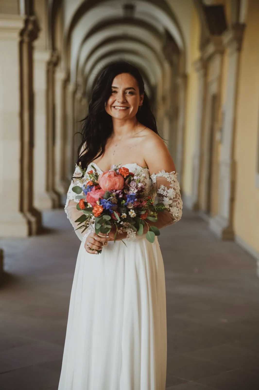 Caro & Dominik – Eine lächelnde Braut im weißen Kleid steht mit einem bunten Blumenstrauß in einem Säulengang