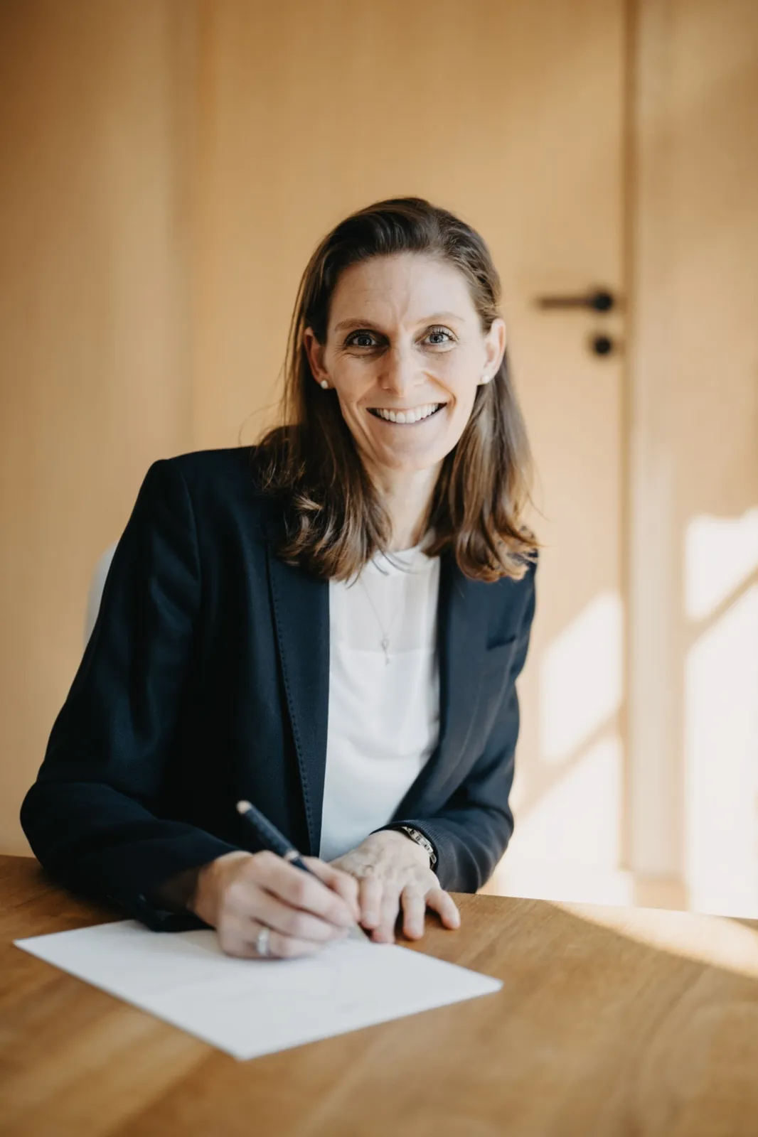 Franziska Hildenbrandt – Eine lächelnde Frau im Business-Outfit sitzt an einem Holztisch und schreibt auf Papier