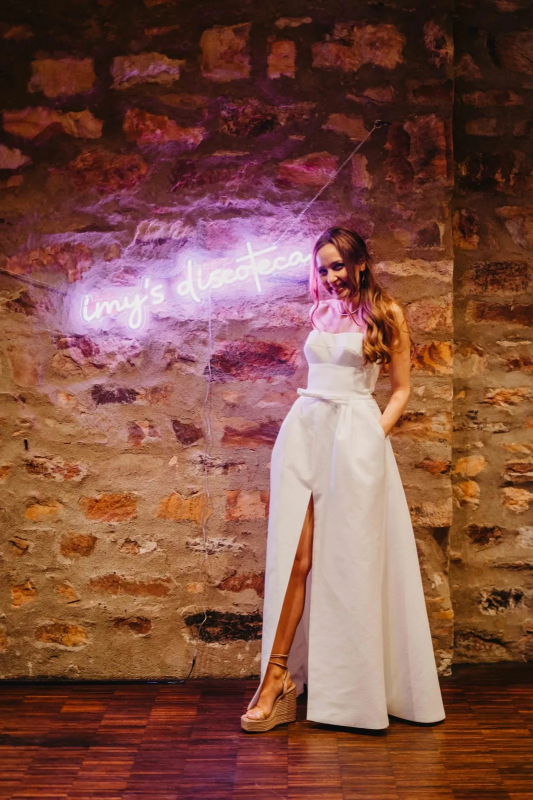 Imy & Felix – Eine lächelnde Frau im weißen Kleid steht vor einer Steinwand mit einem Neon-Schild
