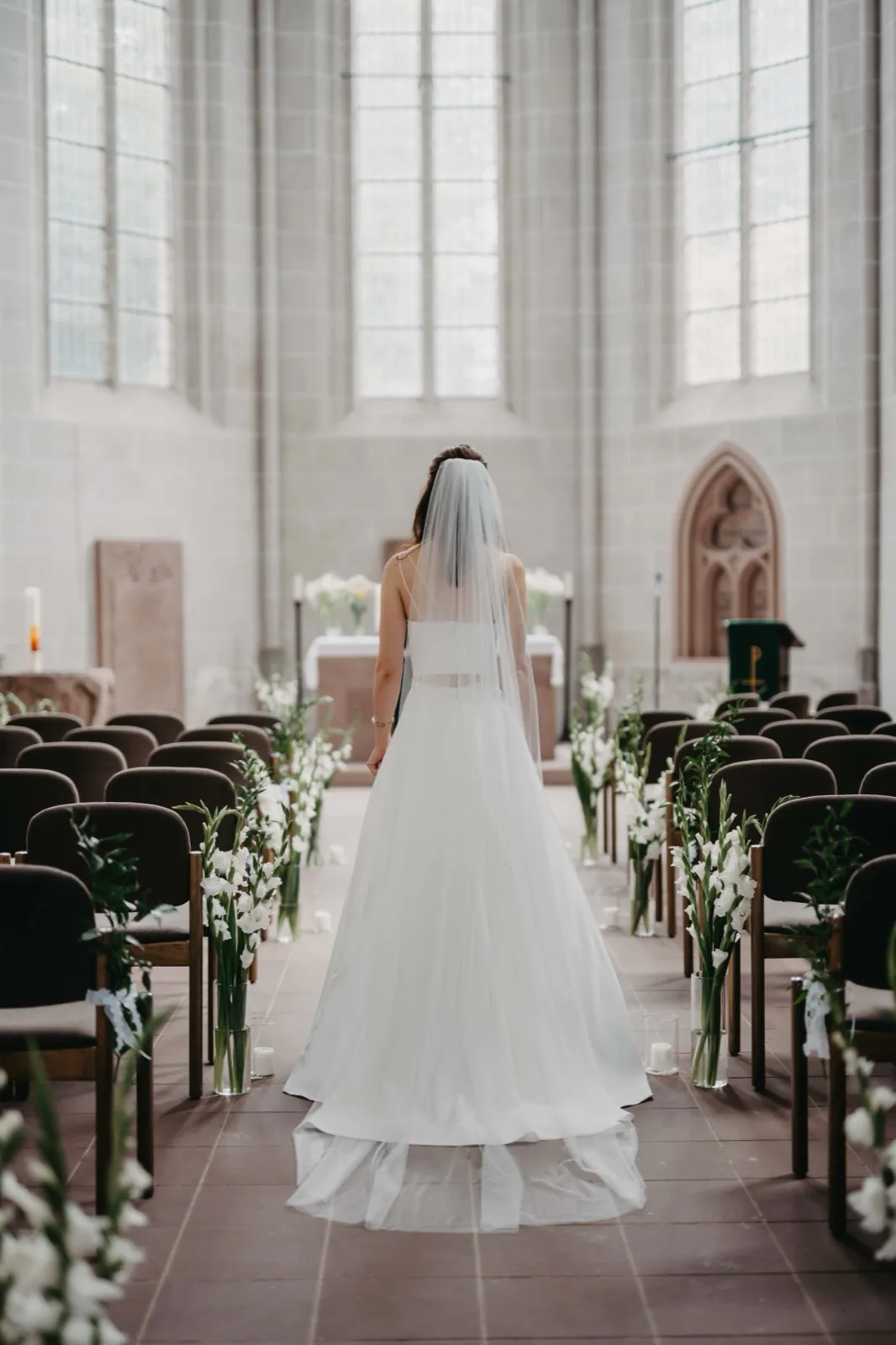 Isa & Nico – Eine Braut von hinten im weißen Kleid steht im Mittelgang einer hellen Kirche