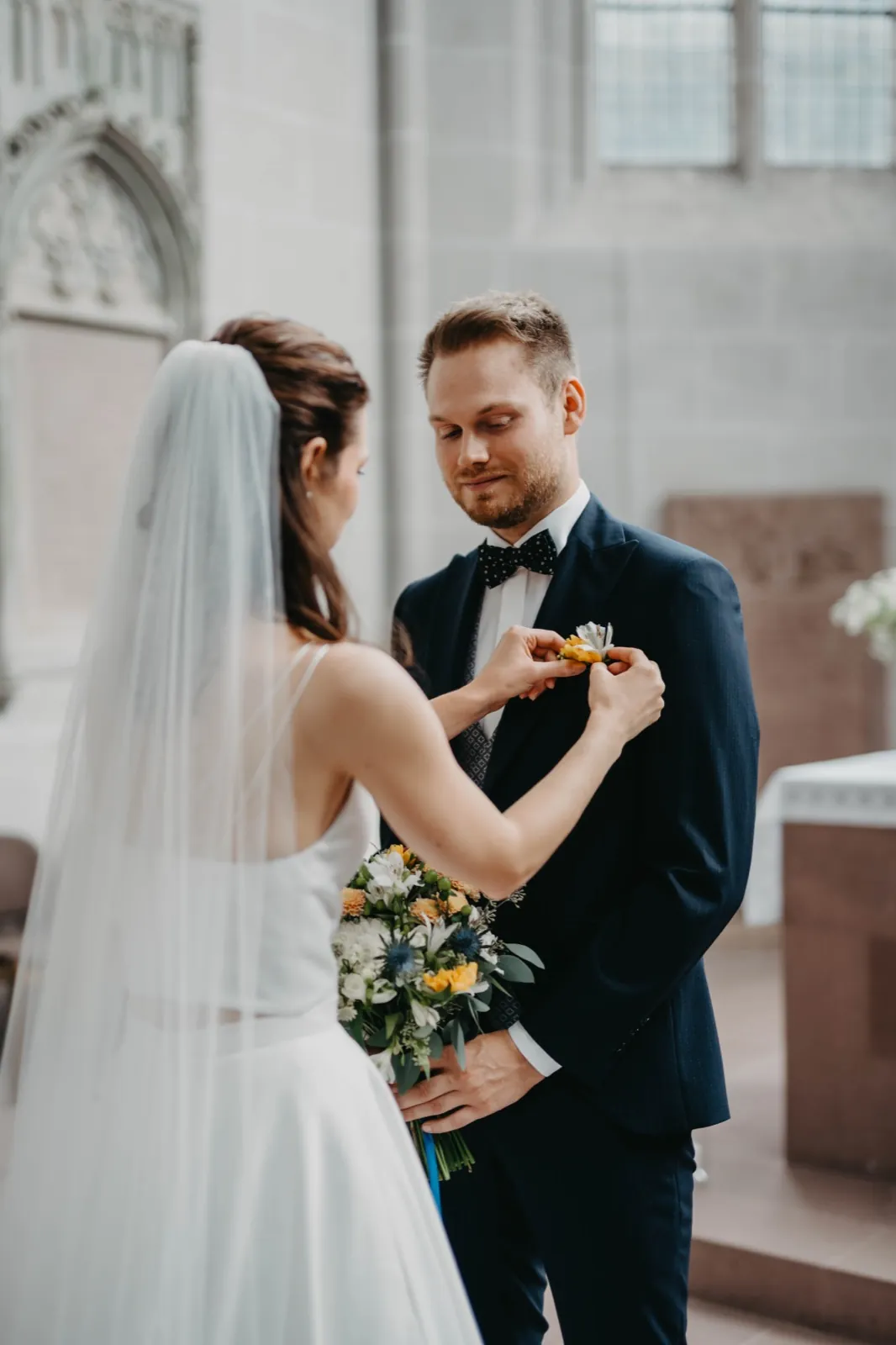 Isa & Nico – Die Braut befestigt eine Ansteckblume am Revers des Bräutigams in einer Kirche