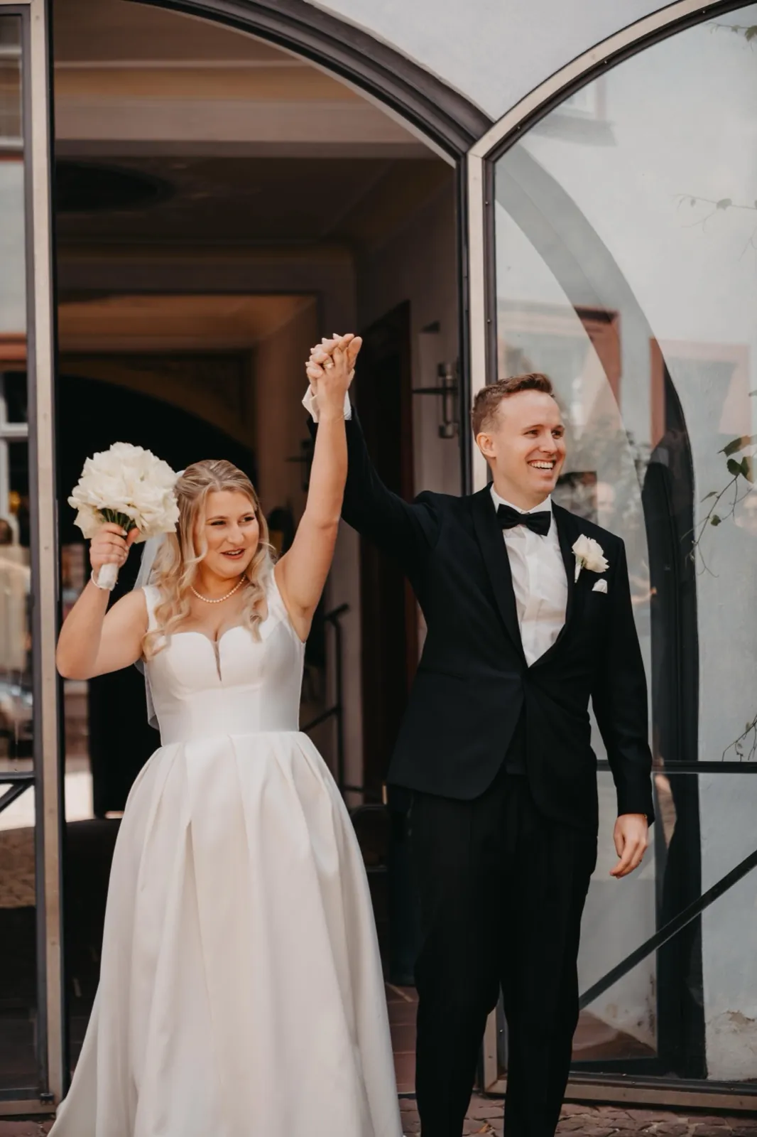 Janina & Alexander – Ein frisch verheiratetes Paar verlässt lächelnd das Gebäude mit erhobenen Händen