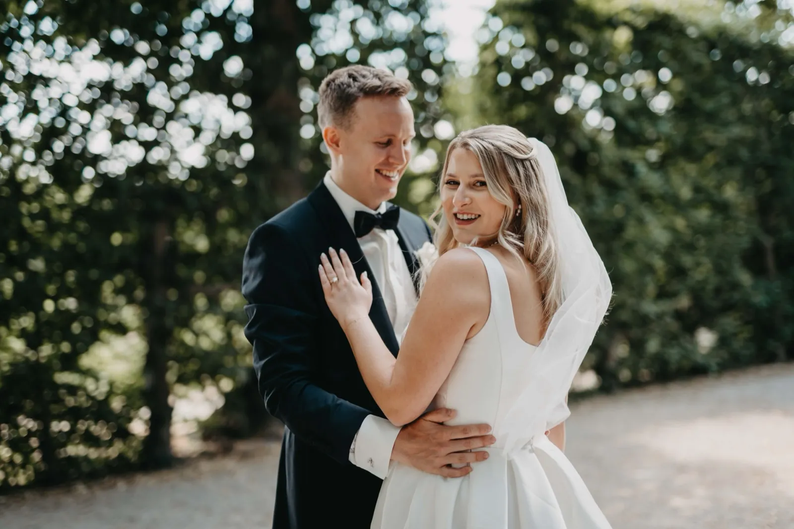 Janina & Alexander – Ein lächelndes Brautpaar umarmt sich im Freien vor grünem Hintergrund