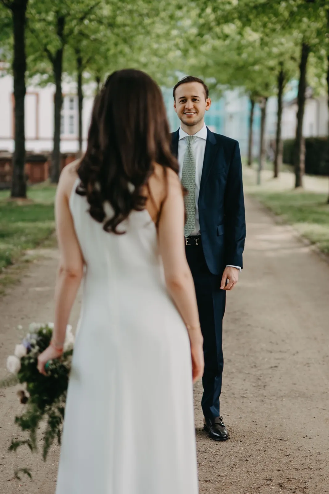 Jessica & Adrian – Ein Bräutigam lächelt seine Braut an, die ihm auf einem von Bäumen gesäumten Weg gegenübersteht