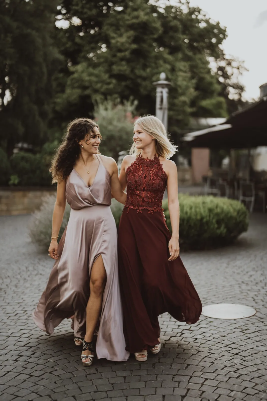 Julia & Chris – Zwei elegant gekleidete Frauen gehen lachend Arm in Arm über ein Kopfsteinpflaster