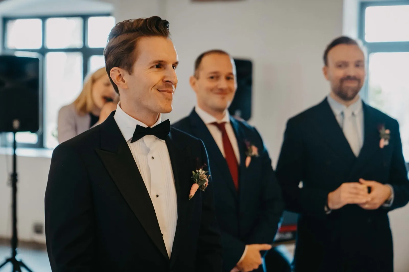Julia & Florian – Der Bräutigam im Smoking blickt lächelnd zur Seite, während seine Trauzeugen hinter ihm stehen