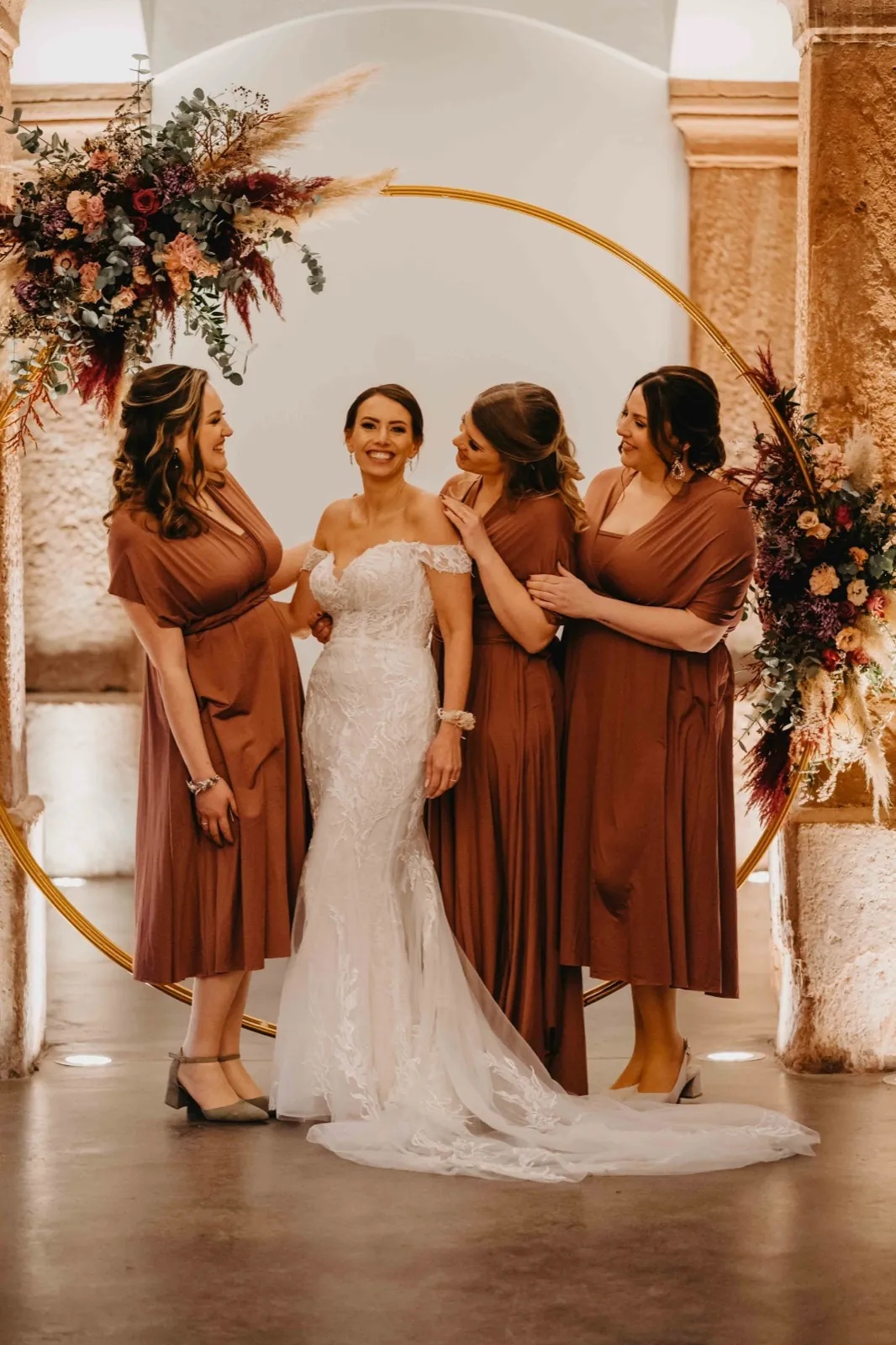 Julia & Florian – Eine Braut im weißen Hochzeitskleid steht lächelnd mit ihren drei Brautjungfern vor einem Bogen