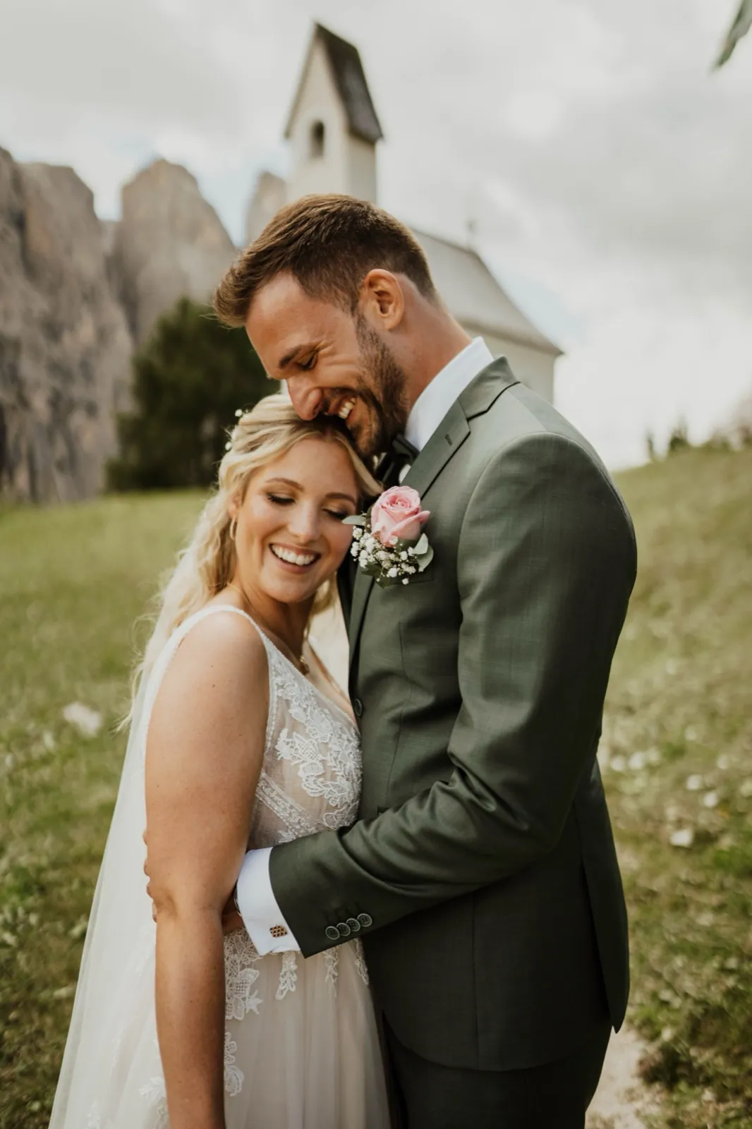 Katrin & Markus – Ein lächelndes Brautpaar umarmt sich innig vor einer Berglandschaft mit einer kleinen Kapelle