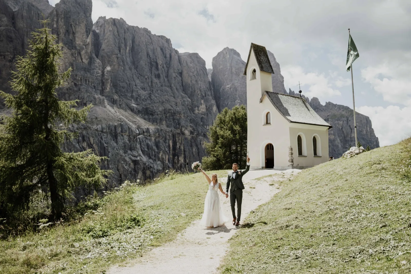 Katrin & Markus – Ein Brautpaar läuft Hand in Hand einen Pfad hinunter vor einer kleinen Bergkapelle
