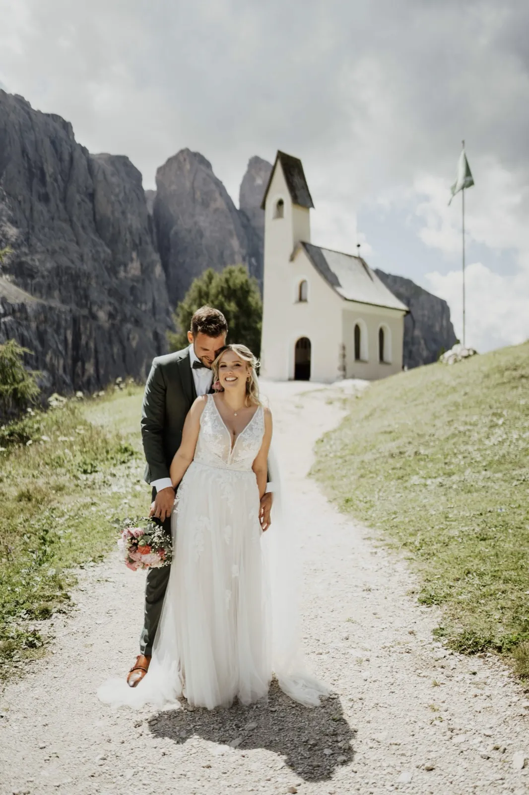 Katrin & Markus – Ein Brautpaar steht lächelnd auf einem Schotterweg vor einer kleinen Kapelle in den Bergen