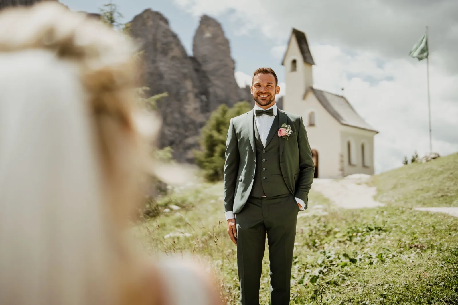 Katrin & Markus – Ein Bräutigam im grünen Anzug blickt lächelnd zur Braut vor einer kleinen Bergkapelle