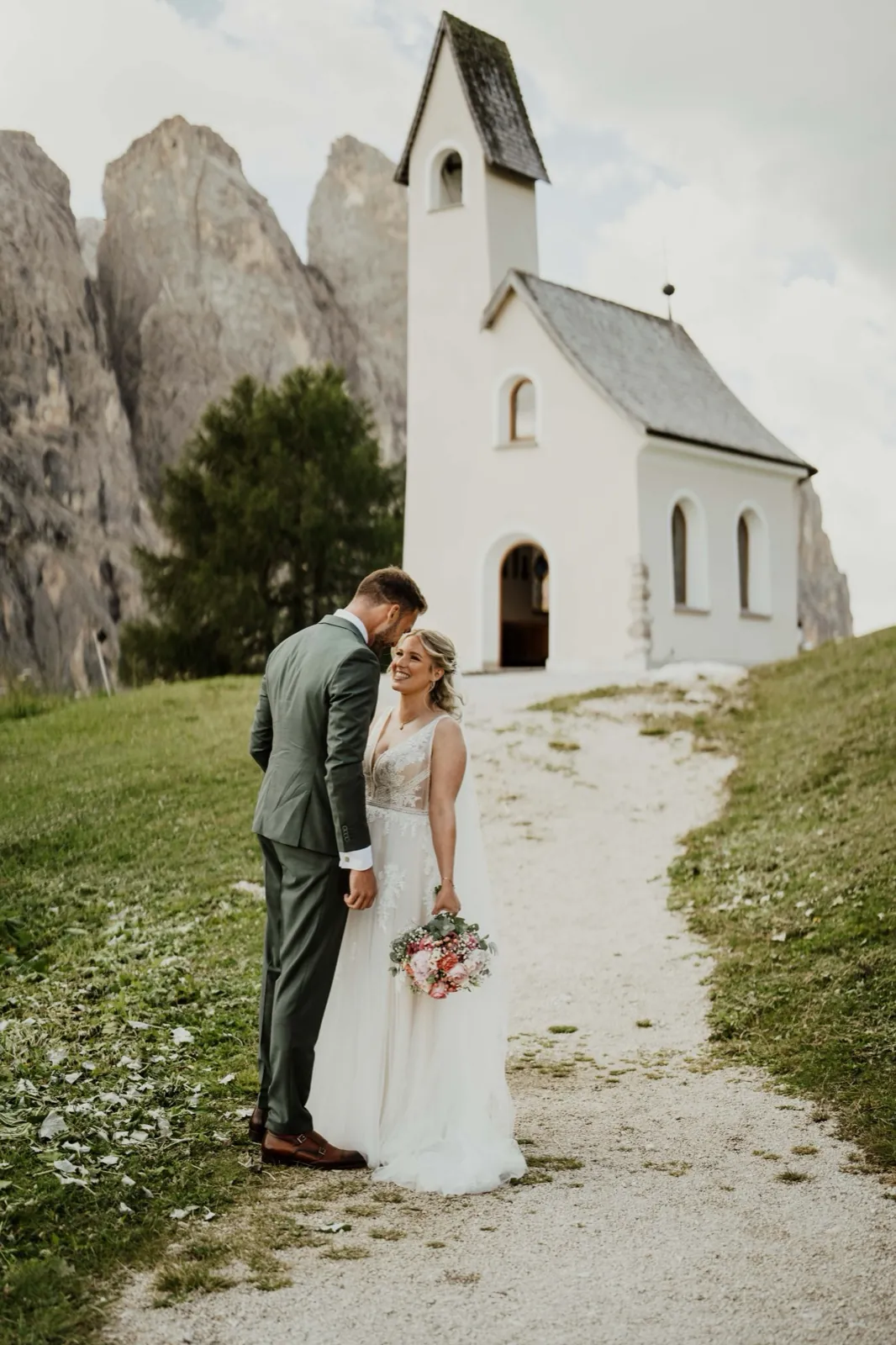 Katrin & Markus – Ein Brautpaar steht lächelnd auf einem Pfad vor einer kleinen Kapelle in den Bergen