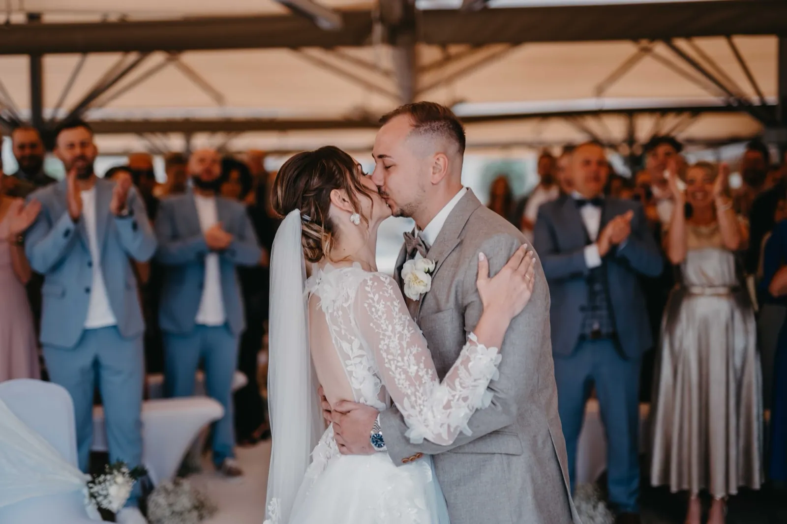 Kristina & Pascal – Das Brautpaar küsst sich während der Hochzeitszeremonie, während die Gäste im Hintergrund applaudieren