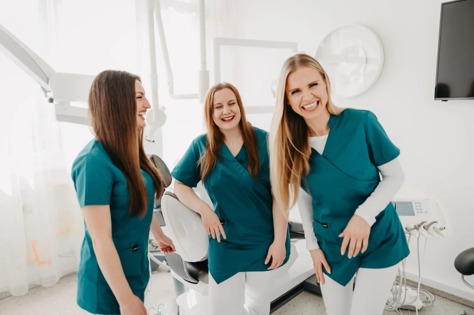 May – Drei Frauen in türkisfarbener Arbeitskleidung lachen in einer modernen Zahnarztpraxis
