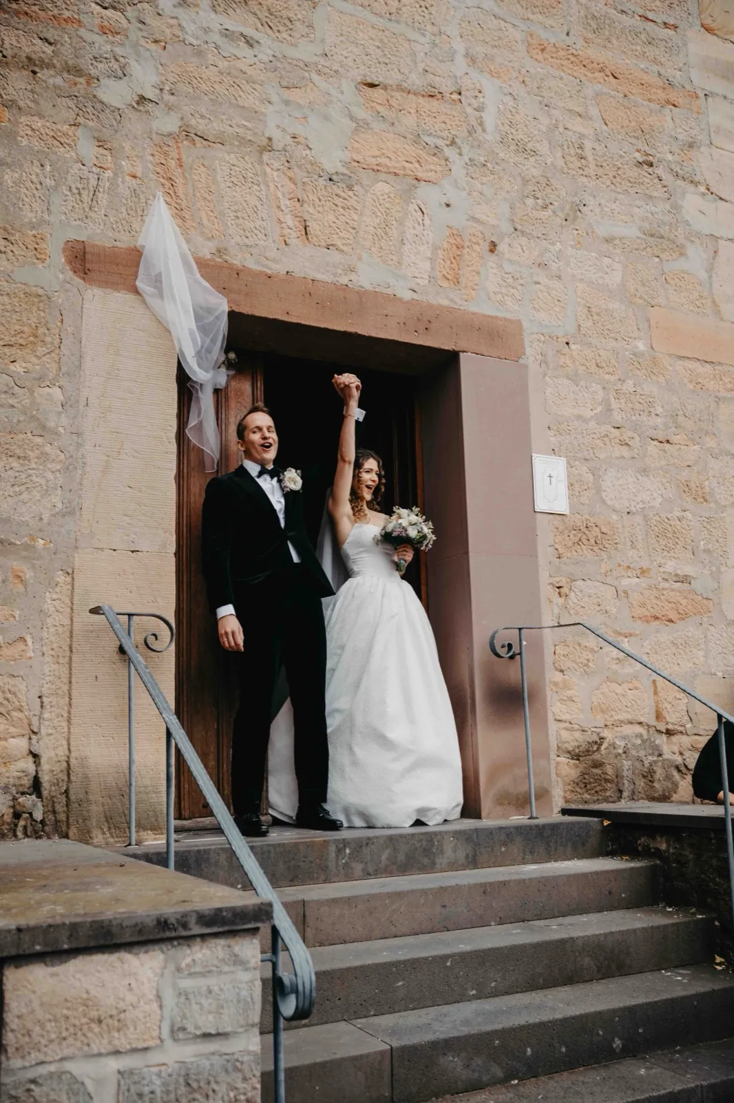 Mia & Clemens – Ein frisch verheiratetes Paar verlässt jubelnd die Kirche über eine Steintreppe
