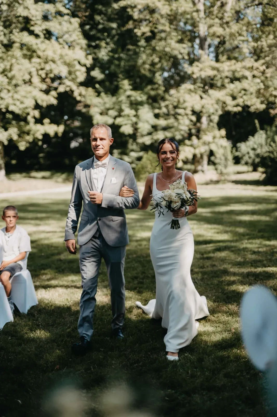 Michelle & Felix – Eine Braut läuft in Begleitung eines Mannes im grauen Anzug über eine Wiese