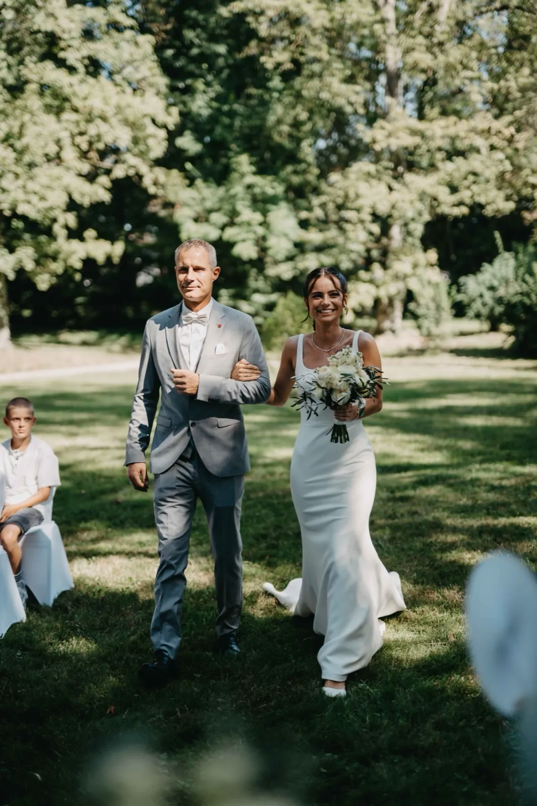 Michelle & Felix – Eine Braut läuft in Begleitung eines Mannes im grauen Anzug über eine Wiese