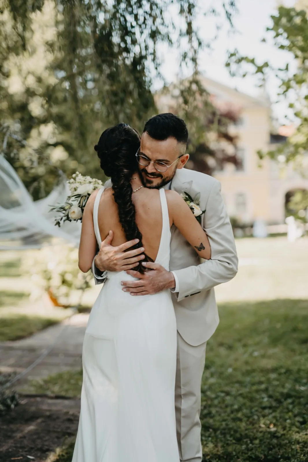 Michelle & Felix – Ein Bräutigam mit Brille umarmt seine Braut im weißen Kleid im Freien