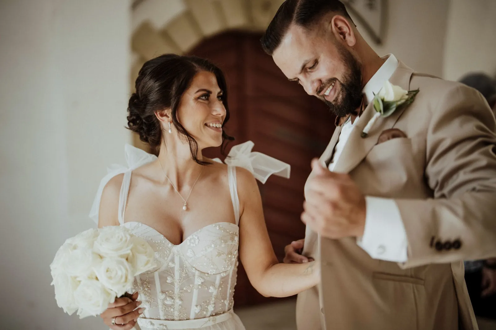 Nadine & Raphael – Ein lächelndes Brautpaar schaut sich an, während die Braut einen weißen Rosenstrauß hält