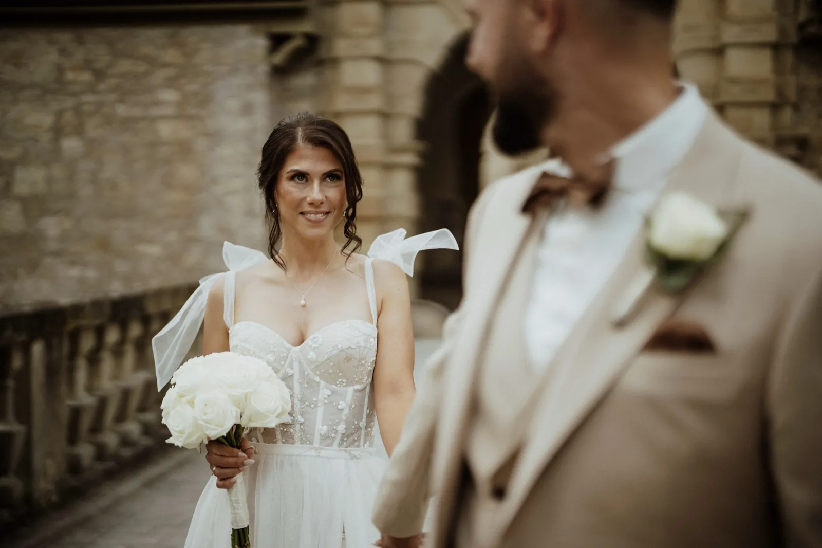 Nadine & Raphael – Eine Braut blickt lächelnd ihren Bräutigam an, während sie einen weißen Rosenstrauß hält