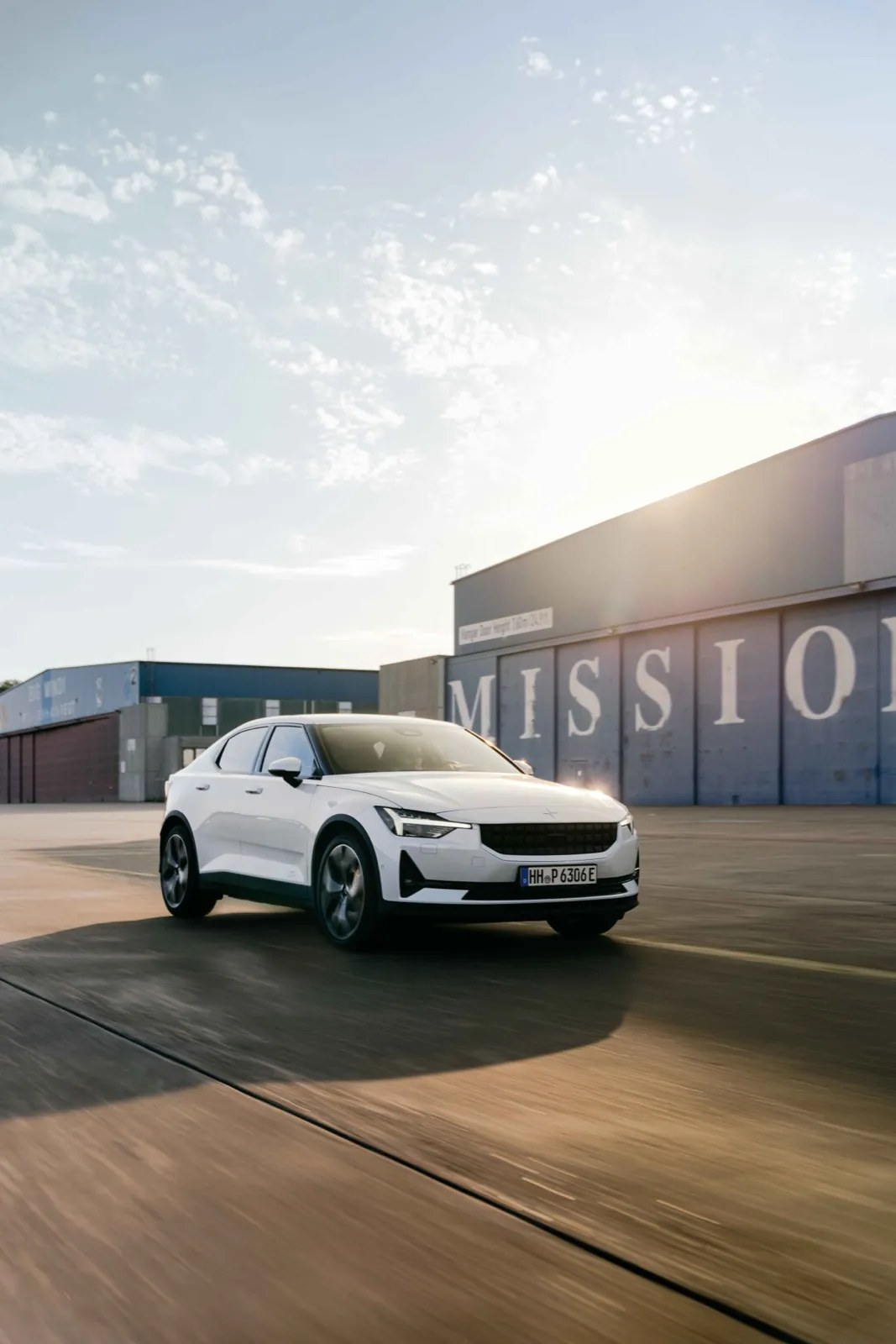 Polestar – Ein weißes Elektroauto fährt bei Sonnenuntergang auf einem Flugplatzgelände vor einem blauen Hangar