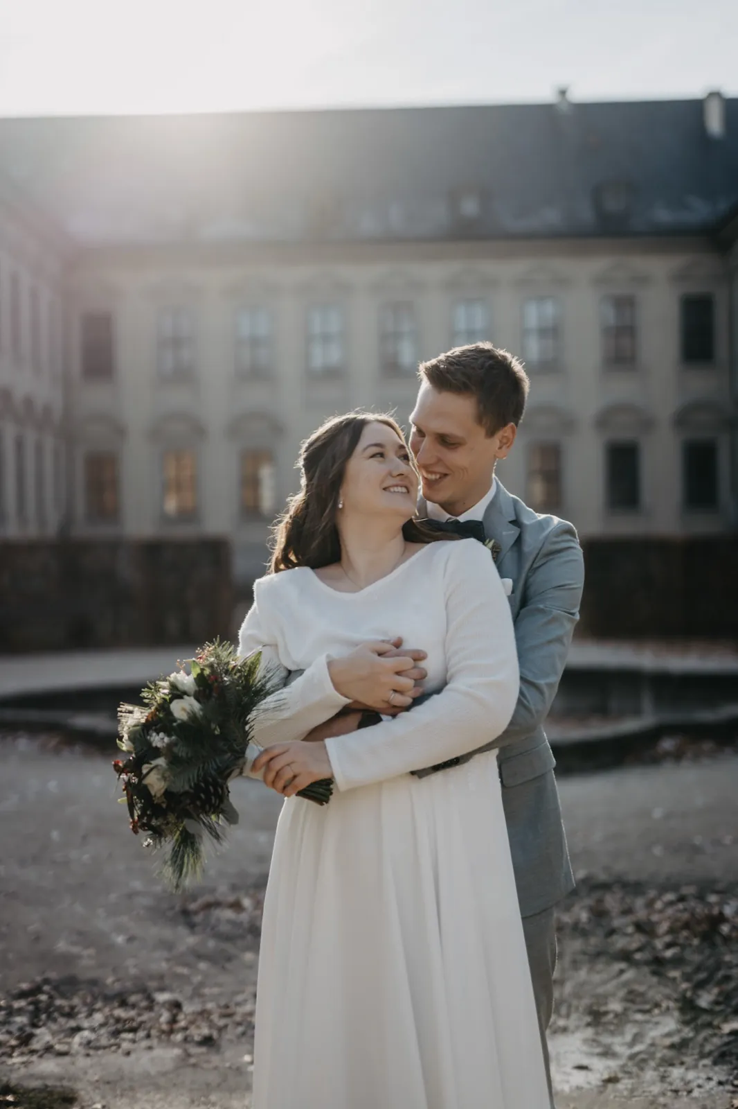 Sophie & Mirko – Ein Brautpaar umarmt sich lächelnd im Freien vor einem historischen Gebäude