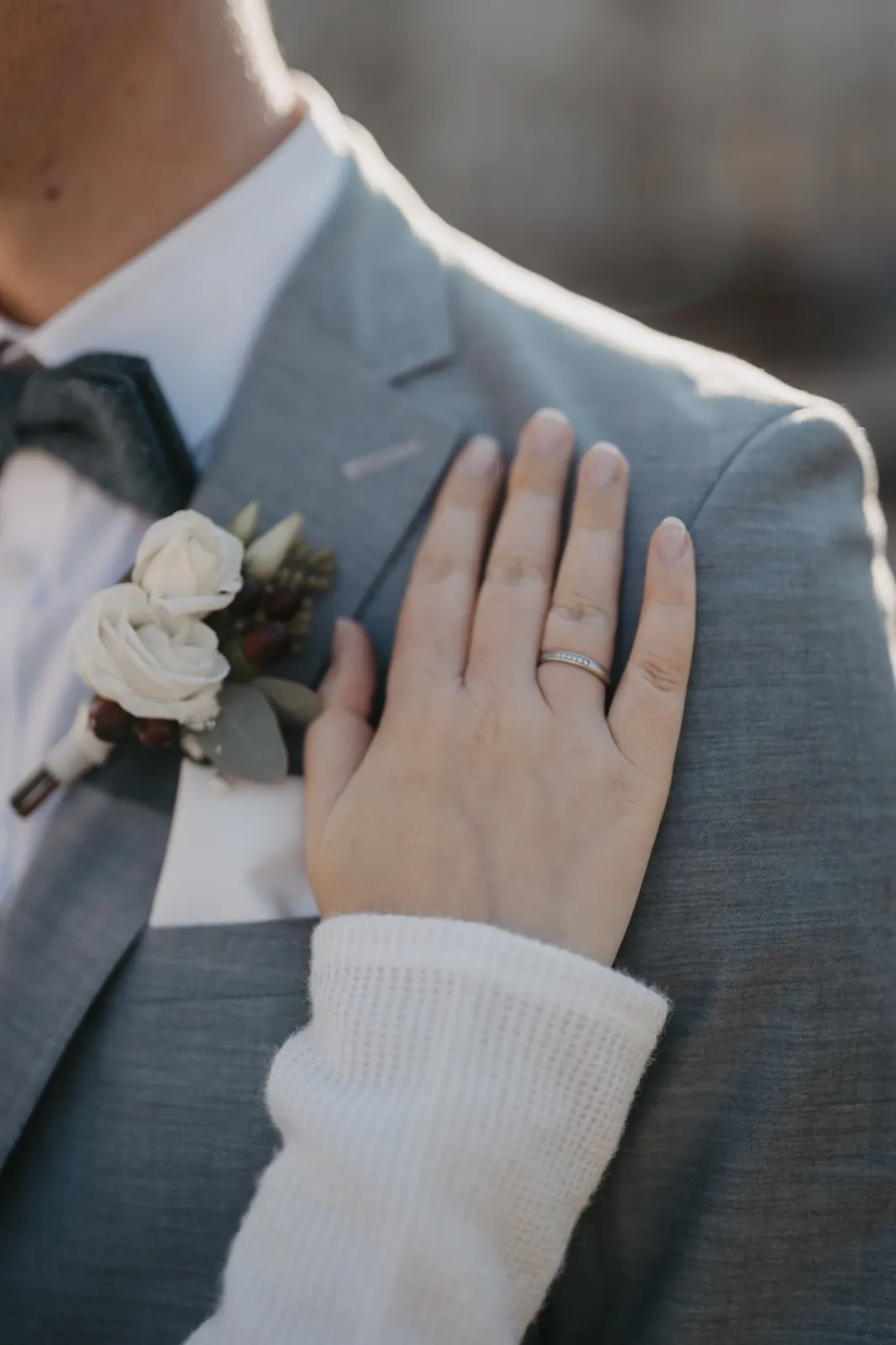 Sophie & Mirko – Eine Hand mit einem Ehering liegt auf der Schulter eines Mannes im grauen Anzug