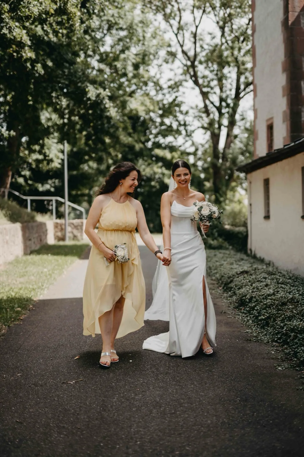 Verena & Rafael – Eine Braut und eine Frau im gelben Kleid gehen Hand in Hand einen Weg entlang