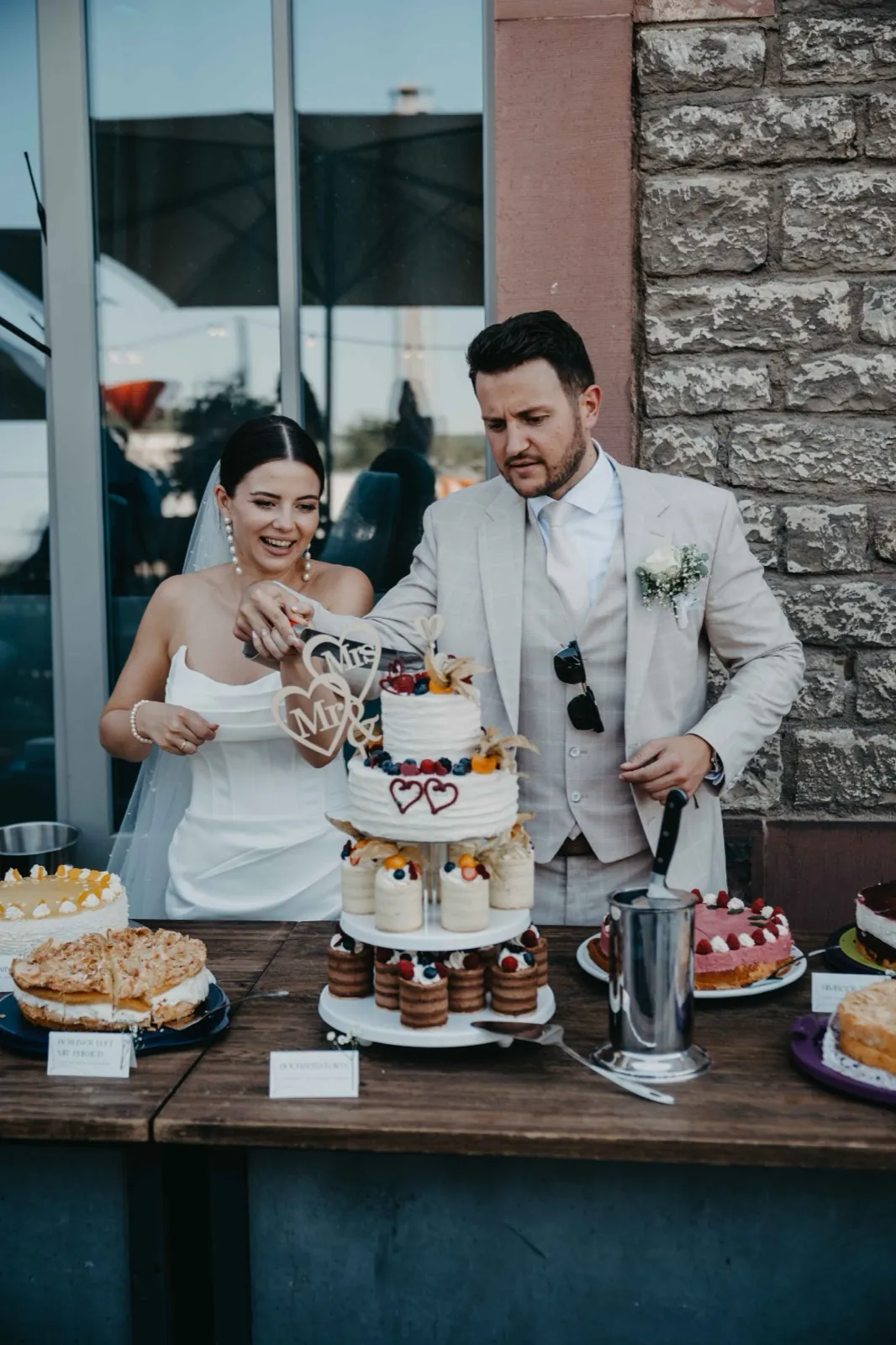 Verena & Rafael – Ein Brautpaar schneidet gemeinsam die mehrstöckige Hochzeitstorte an einem Tisch an