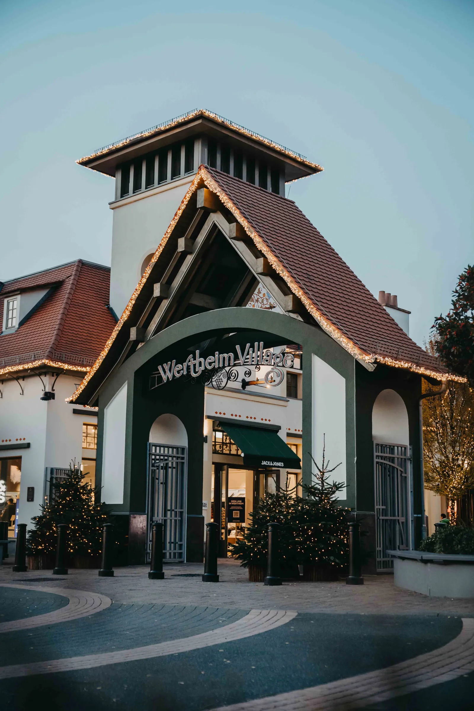 Wertheim Village – Der beleuchtete Eingangsbereich des Wertheim Village mit weihnachtlicher Dekoration und Tannenbäumen